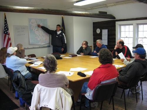 a group of people sitting around a large conference table and looking at someone pointing to a map. Photo by Tara Mastel, MSU Extension.