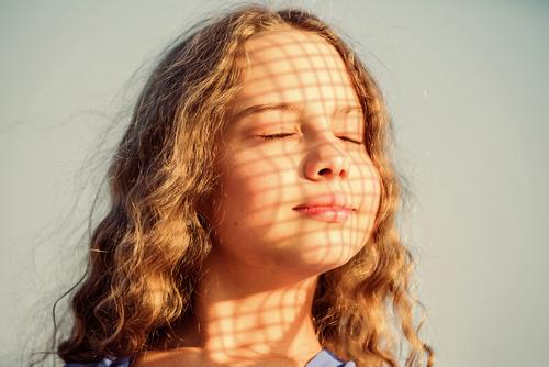 A young girl with brown hair closes her eyes in sunlight through a window.