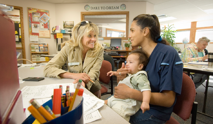 A woman sites at a table with a mother holding a young infant. 