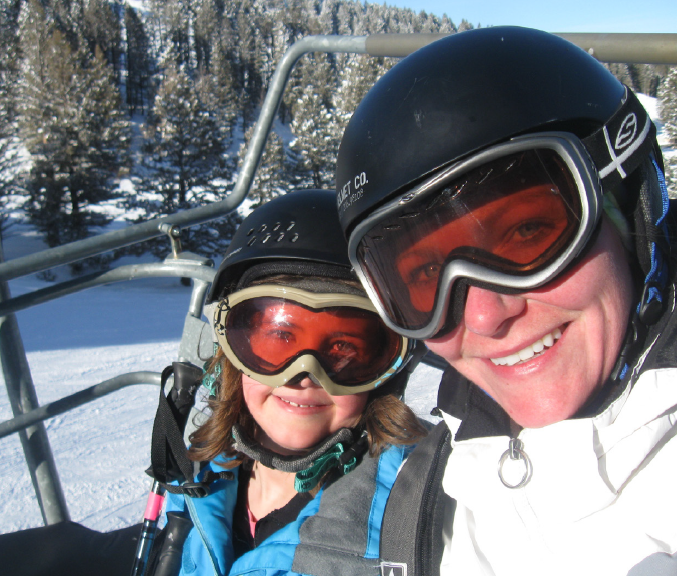 A woman and a young girl smile at the camera as they ride a chairlift at a ski hill