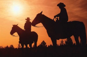 Two silhouetted cowboys riding horses in the sunset